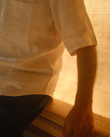 Close up of the pocket detail of an off-white linen shirt worn by a man sitting on a bamboo rug