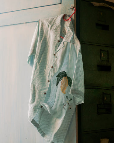 Green and white stripes linen shirt with embroidered mangoes hung on a blue door.