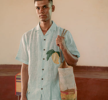 a man standing in a yoga hall wearing co-ordinated green and white stripe linen shirt and shorts with embroidered mangoes. Front view