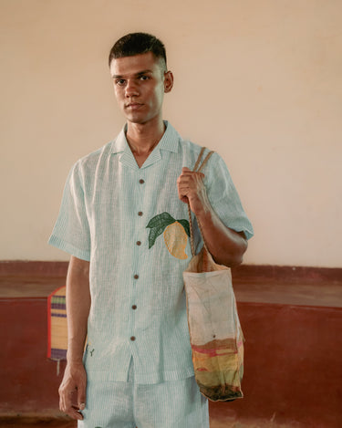 a man standing in a yoga hall wearing co-ordinated green and white stripe linen shirt and shorts with embroidered mangoes. Front view