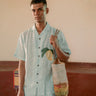 a man standing in a yoga hall wearing co-ordinated green and white stripe linen shirt and shorts with embroidered mangoes. Front view