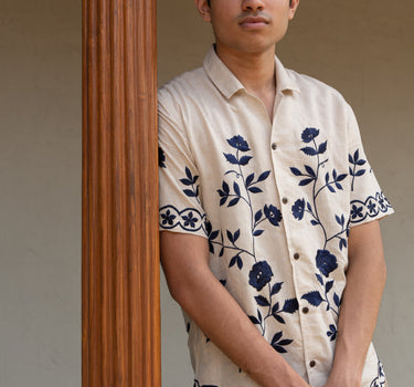 a man wearing beige linen blue floral hand-embroidered shirt leaning against a pillar.