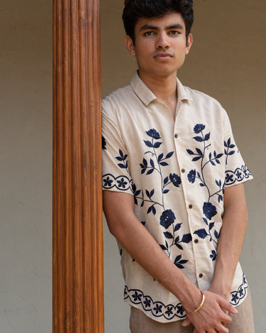 a man wearing beige linen blue floral hand-embroidered shirt leaning against a pillar.