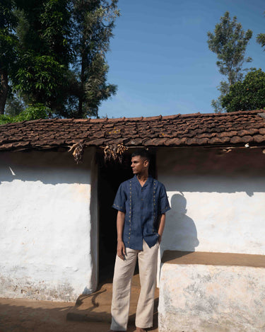 A man in eco-friendly, buds in blue hand-embroidered linen shirt in front of a hut. half sleeves shirt, Material: linen, front view